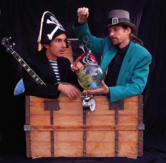 Zwei Männer in Kostümen, einer als Pirat mit Banjo, der andere mit blaugrüner Jacke und grauem Hut, stehen hinter einer offenen Holztruhe mit einem Fischglas und einer Taschenuhr.