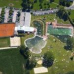 Luftaufnahme einer Freizeitanlage mit Tennisplätzen, Schwimmbädern, einem Parkplatz und Grünflächen, die von Straßen und Bäumen umgeben sind.