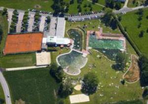 Luftaufnahme einer Freizeitanlage mit Tennisplätzen, Schwimmbädern, einem Parkplatz und Grünflächen, die von Straßen und Bäumen umgeben sind.