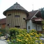 Ein Rundbau mit Strohdach steht neben einem Haus mit rotem Ziegeldach, umgeben von Grünpflanzen und gelben Blumen.