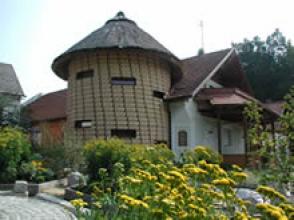 Ein Rundbau mit Strohdach steht neben einem Haus mit rotem Ziegeldach, umgeben von Grünpflanzen und gelben Blumen.