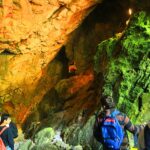 Eine Gruppe von Menschen mit Rucksäcken erkundet eine Höhle mit felsigen Wänden, orangefarbener Beleuchtung und grünen, moosbewachsenen Oberflächen.