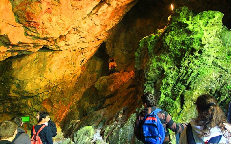 Eine Gruppe von Menschen mit Rucksäcken erkundet eine Höhle mit felsigen Wänden, orangefarbener Beleuchtung und grünen, moosbewachsenen Oberflächen.