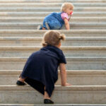 Zwei Kleinkinder krabbeln eine breite Betontreppe hinauf, wobei jedes von ihnen ein anderes farbiges Outfit trägt.