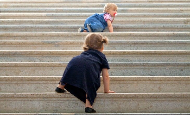 Zwei Kleinkinder krabbeln eine breite Betontreppe hinauf, wobei jedes von ihnen ein anderes farbiges Outfit trägt.