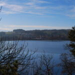 Ein ruhiger See mit kahlen Bäumen im Vordergrund, einer kleinen Stadt und Hügeln im Hintergrund unter einem blauen Himmel mit leichten Wolken.