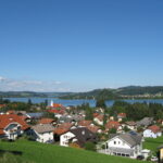 Eine kleine Stadt mit Häusern und roten Dächern liegt in der Nähe eines Sees, mit grünen Hügeln und blauem Himmel im Hintergrund.