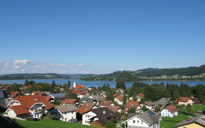 Eine kleine Stadt mit Häusern und roten Dächern liegt in der Nähe eines Sees, mit grünen Hügeln und blauem Himmel im Hintergrund.