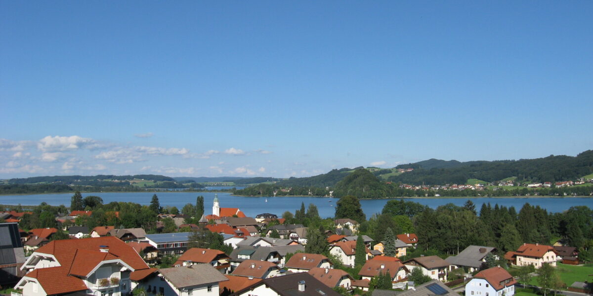 Eine kleine Stadt mit roten Dächern liegt in der Nähe eines Sees, umgeben von grünen Hügeln unter einem klaren blauen Himmel.