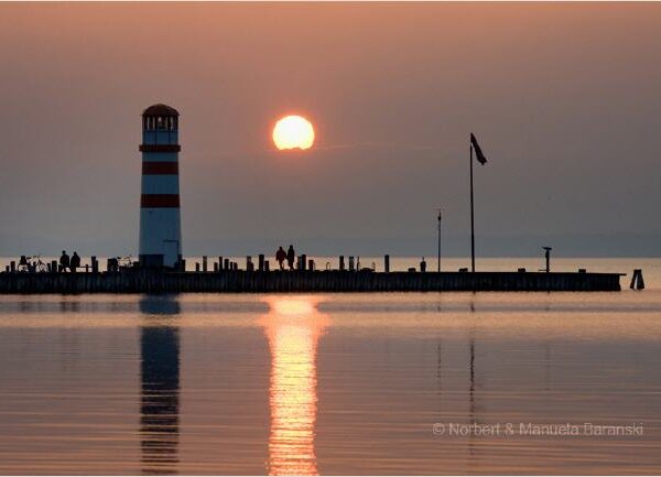 Ein gestreifter Leuchtturm und Menschen auf einem Pier heben sich von einem ruhigen See bei Sonnenuntergang ab, wobei sich die Sonne auf dem Wasser spiegelt.