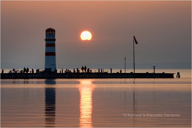 Ein gestreifter Leuchtturm und Menschen auf einem Pier heben sich von einem ruhigen See bei Sonnenuntergang ab, wobei sich die Sonne auf dem Wasser spiegelt.
