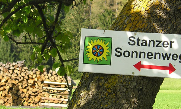 Ein Schild mit der Aufschrift "Stanzer Sonnenweg" mit einem Sonnensymbol und einem roten Pfeil ist an einem Baum auf einer Wiese in der Nähe von gestapeltem Brennholz angebracht.