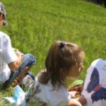 Drei Kinder in weißen T-Shirts mit bunten Schmetterlingsaufdrucken sitzen im Gras in einem sonnigen Außenbereich.