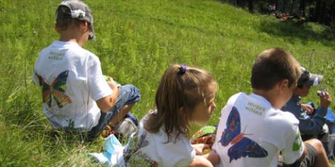 Drei Kinder in weißen T-Shirts mit bunten Schmetterlingsaufdrucken sitzen im Gras in einem sonnigen Außenbereich.