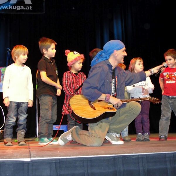 Ein Mann mit einer Gitarre kniet mit mehreren Kindern auf der Bühne und hält einem kleinen Jungen ein Mikrofon hin, während andere in der Nähe stehen.