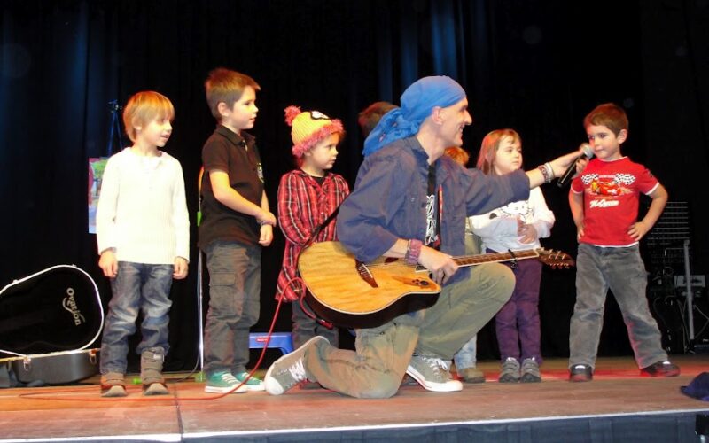 Ein Mann mit einer Gitarre kniet mit mehreren Kindern auf der Bühne und hält einem kleinen Jungen ein Mikrofon hin, während andere in der Nähe stehen.