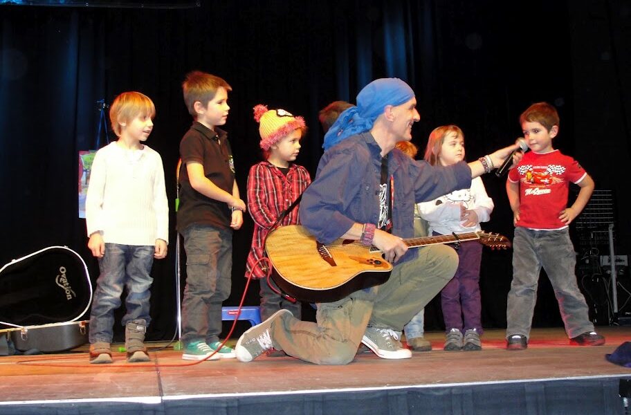 Ein Mann mit einer Gitarre kniet mit mehreren Kindern auf der Bühne und hält einem kleinen Jungen ein Mikrofon hin, während andere in der Nähe stehen.