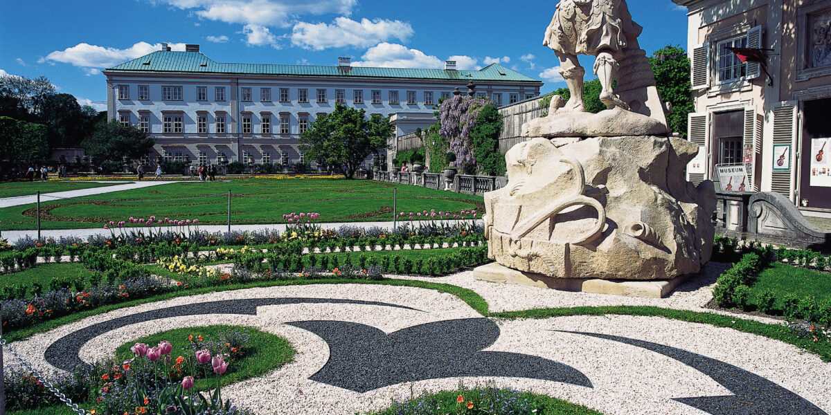 Ein formaler Garten mit dekorativ gemusterten Blumenbeeten, einer großen Steinstatue und einem historischen Gebäude im Hintergrund unter blauem Himmel.