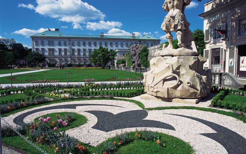 Ein formaler Garten mit dekorativ gemusterten Blumenbeeten, einer großen Steinstatue und einem historischen Gebäude im Hintergrund unter blauem Himmel.