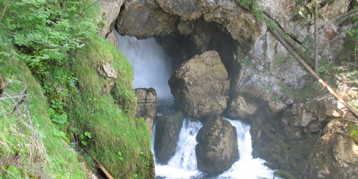 Ein Wasserfall fließt durch eine felsige Höhle, die von grüner Vegetation und moosbewachsenen Felsen in einem Waldgebiet umgeben ist.