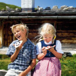 Zwei Kinder sitzen vor einer rustikalen Holzhütte und essen verzierte Kekse. Der Junge lacht mit offenem Mund, während das Mädchen auf ihre Leckerei hinunterschaut. Im Hintergrund sind grüne Hügel zu sehen.