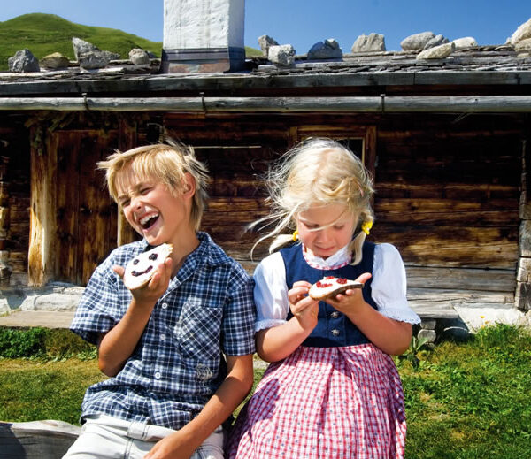 Zwei Kinder sitzen vor einer rustikalen Holzhütte und essen verzierte Kekse. Der Junge lacht mit offenem Mund, während das Mädchen auf ihre Leckerei hinunterschaut. Im Hintergrund sind grüne Hügel zu sehen.