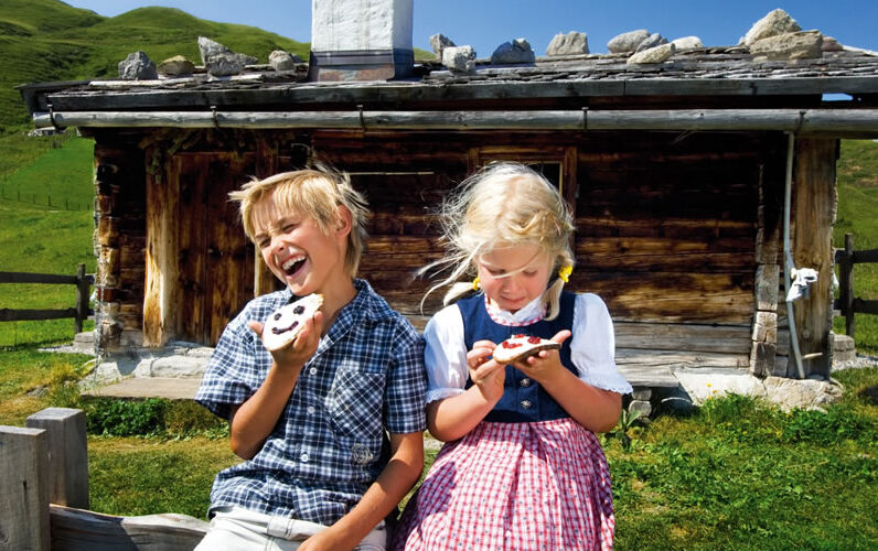 Zwei Kinder sitzen vor einer rustikalen Holzhütte und essen verzierte Kekse. Der Junge lacht mit offenem Mund, während das Mädchen auf ihre Leckerei hinunterschaut. Im Hintergrund sind grüne Hügel zu sehen.