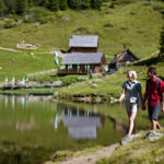 Zwei Personen spazieren am Rande eines ruhigen Sees entlang, mit grünen Hügeln und Holzhütten im Hintergrund.
