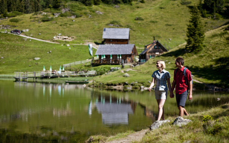 Zwei Personen spazieren am Rande eines ruhigen Sees entlang, mit grünen Hügeln und Holzhütten im Hintergrund.