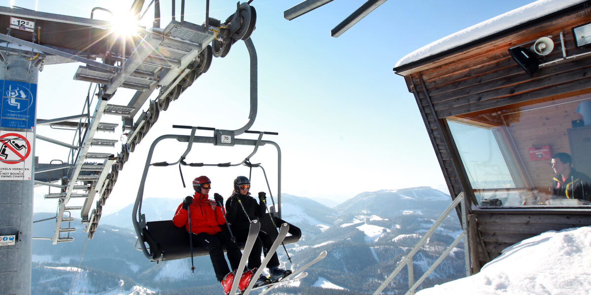 Zwei Skifahrer fahren mit einem Sessellift über verschneite Berge, vorbei an einer hölzernen Kabine des Liftbetreibers und unter strahlend blauem Himmel.