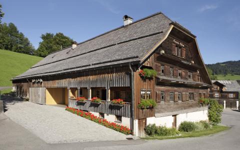 Ein großes traditionelles Bauernhaus aus Holz mit Blumenkästen unter den Fenstern, das an einer Kopfsteinpflasterstraße in einer ländlichen Gegend mit grünen Hügeln und Bäumen liegt.