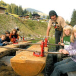 Mehrere Kinder und ein Erwachsener schneiden mit handbetriebenen Sägen Holzstämme im Freien in einer ländlichen, bergigen Gegend mit Bäumen und Holzgebäuden im Hintergrund.