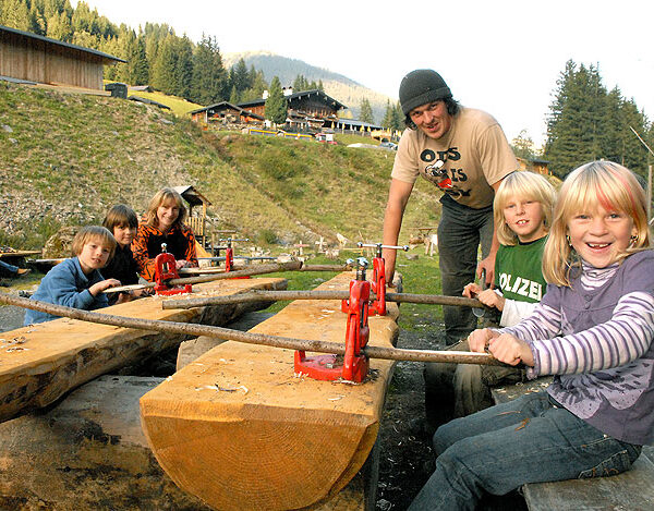 Mehrere Kinder und ein Erwachsener schneiden mit handbetriebenen Sägen Holzstämme im Freien in einer ländlichen, bergigen Gegend mit Bäumen und Holzgebäuden im Hintergrund.