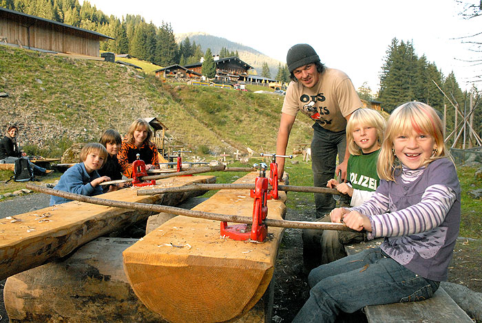 Mehrere Kinder und ein Erwachsener schneiden mit handbetriebenen Sägen Holzstämme im Freien in einer ländlichen, bergigen Gegend mit Bäumen und Holzgebäuden im Hintergrund.