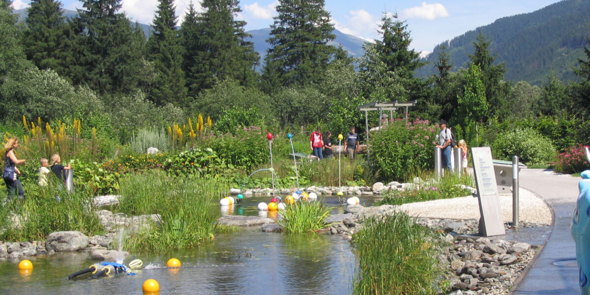 Ein Landschaftsgarten mit einem Teich, schwimmenden Objekten, Menschen, die auf Wegen spazieren gehen, und Bergen und Bäumen im Hintergrund unter einem teilweise bewölkten Himmel.