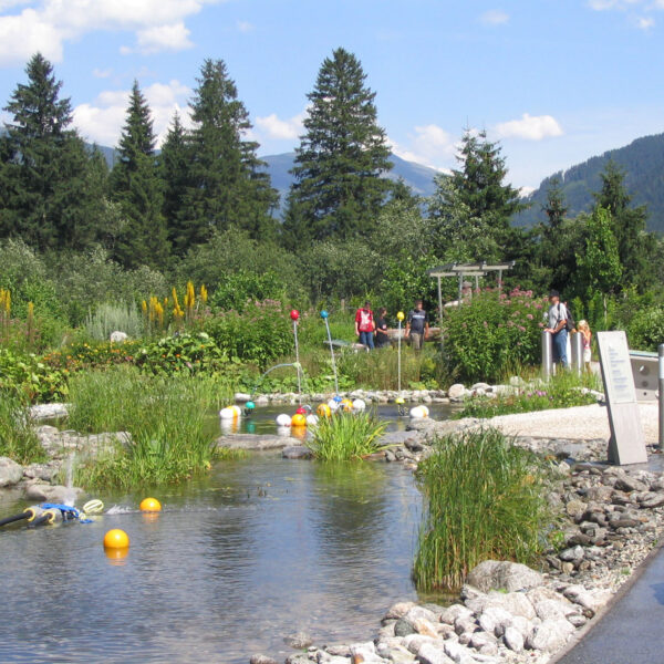 Ein Landschaftsgarten mit einem Teich, schwimmenden Objekten, Menschen, die auf Wegen spazieren gehen, und Bergen und Bäumen im Hintergrund unter einem teilweise bewölkten Himmel.
