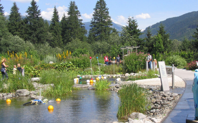 Ein Landschaftsgarten mit einem Teich, schwimmenden Objekten, Menschen, die auf Wegen spazieren gehen, und Bergen und Bäumen im Hintergrund unter einem teilweise bewölkten Himmel.