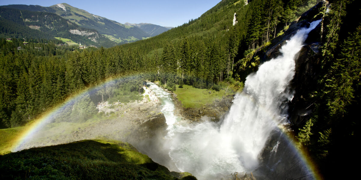 Ein großer Wasserfall stürzt in ein grünes Tal, das von bewaldeten Bergen umgeben ist, und im Nebel ist ein leuchtender Regenbogen zu sehen.