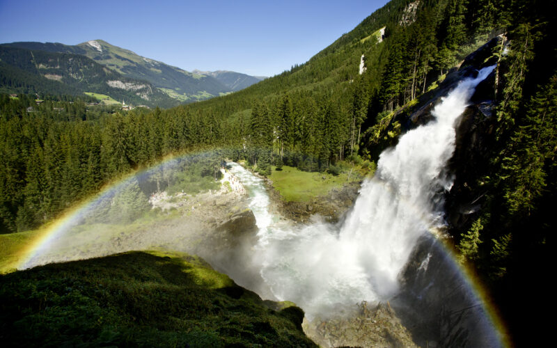 Ein großer Wasserfall stürzt in ein grünes Tal, das von bewaldeten Bergen umgeben ist, und im Nebel ist ein leuchtender Regenbogen zu sehen.