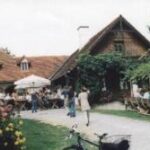 Vor einem rustikalen Gebäude mit Ziegeldach sind Menschen an Tischen im Freien versammelt, umgeben von Pflanzen und Blumen. Im Vordergrund ist ein Fahrrad geparkt.