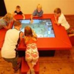 Sechs Kinder interagieren mit einem großen Touchscreen-Tisch in einem Raum mit Holzfußboden, wobei jedes von ihnen auf roten Sitzen um den Tisch herum sitzt oder sich anlehnt.