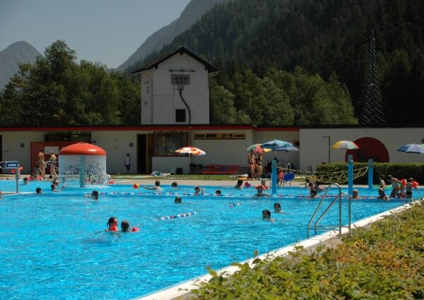 Freibad mit schwimmenden und spielenden Menschen; Berge und Bäume im Hintergrund unter einem klaren Himmel.