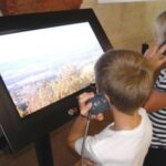 Zwei Kinder stehen vor einem großen interaktiven Bildschirm und halten sich jeweils ein Handgerät ans Ohr, während sie ein Landschaftsbild im Freien auf dem Display betrachten.