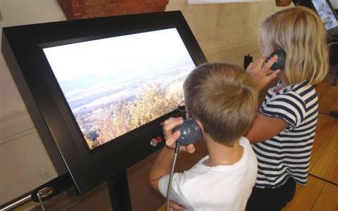 Zwei Kinder stehen vor einem großen interaktiven Bildschirm und halten sich jeweils ein Handgerät ans Ohr, während sie ein Landschaftsbild im Freien auf dem Display betrachten.