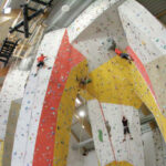 In einer geräumigen Kletterhalle klettern mehrere Personen an hohen Indoor-Felswänden mit verschiedenfarbigen Griffen.