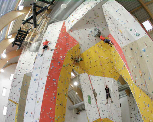 In einer geräumigen Kletterhalle klettern mehrere Personen an hohen Indoor-Felswänden mit verschiedenfarbigen Griffen.