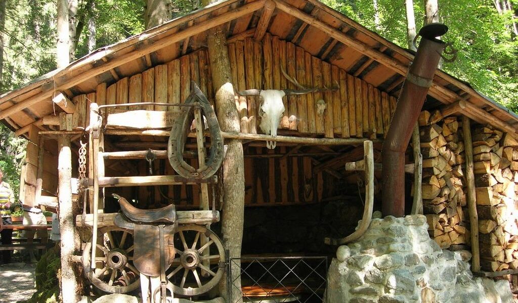 Eine rustikale Holzhütte mit gestapeltem Brennholz, einem montierten Rinderschädel, einem alten Wagenrad und einem Sattel an der Außenwand.