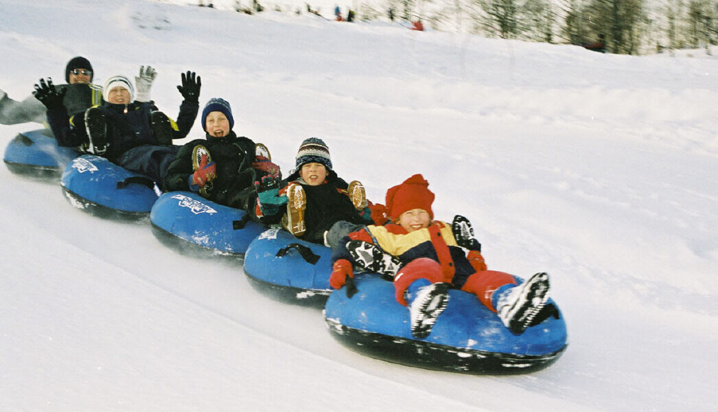 Sechs Personen, darunter Kinder und Erwachsene, fahren gemeinsam in Winterkleidung auf blauen Snowtubes einen verschneiten Hügel hinunter. Im Hintergrund sind Bäume und Schnee zu sehen.