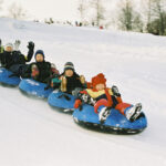Sechs Personen, darunter Kinder und Erwachsene, fahren gemeinsam in Winterkleidung auf blauen Snowtubes einen verschneiten Hügel hinunter. Im Hintergrund sind Bäume und Schnee zu sehen.