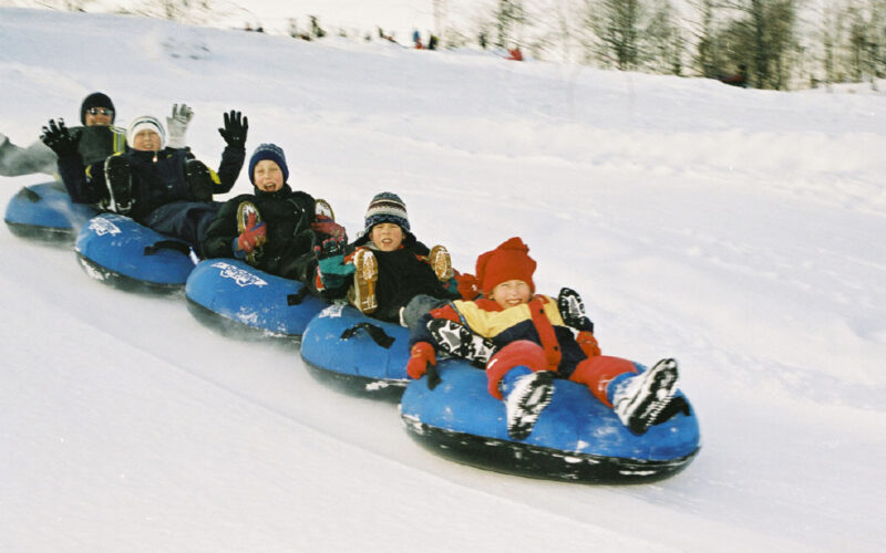 Sechs Personen, darunter Kinder und Erwachsene, fahren gemeinsam in Winterkleidung auf blauen Snowtubes einen verschneiten Hügel hinunter. Im Hintergrund sind Bäume und Schnee zu sehen.